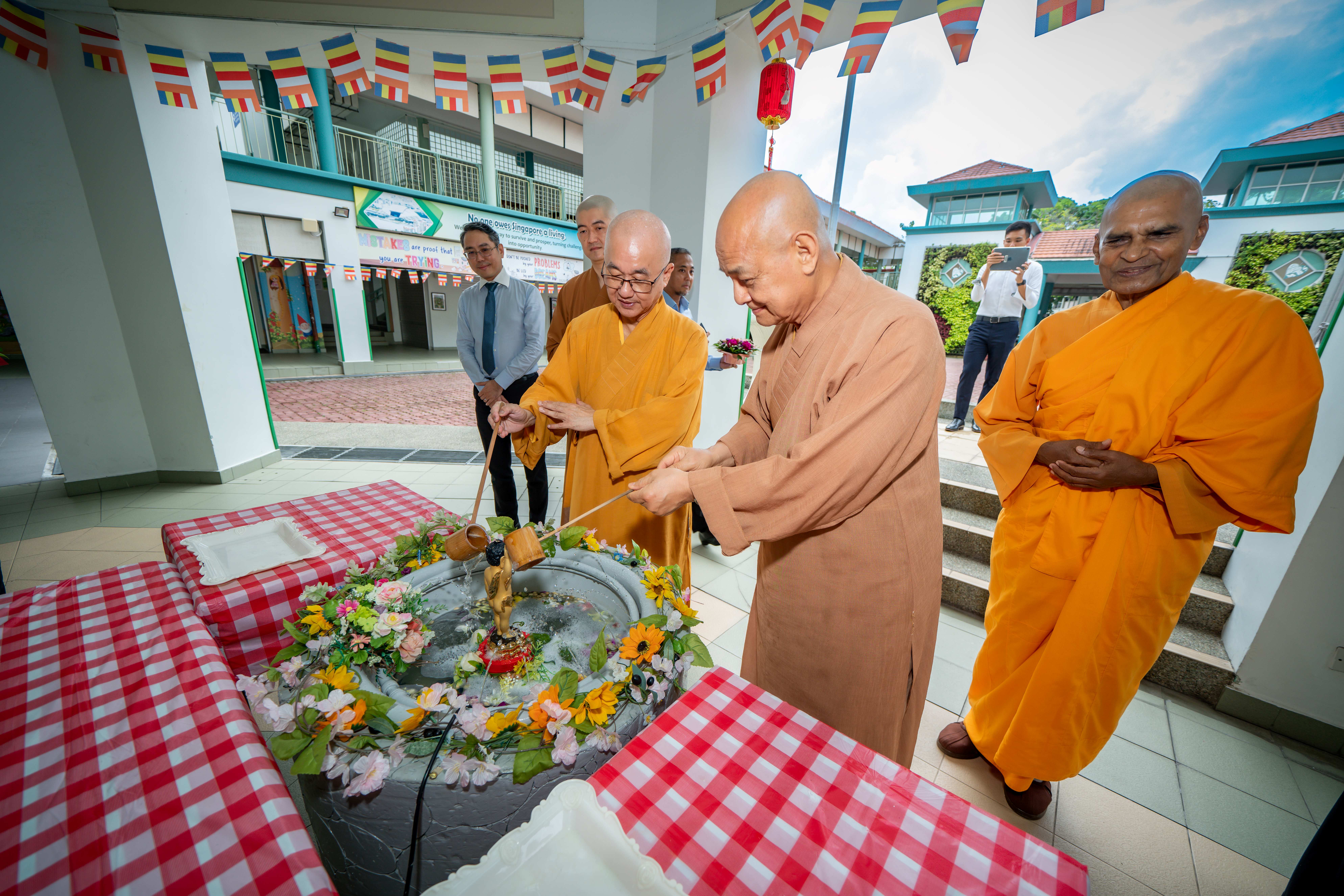 Buddhist monks in orange robes perform a ritual pouring water over a flower-adorned statue outdoors under colorful flags.